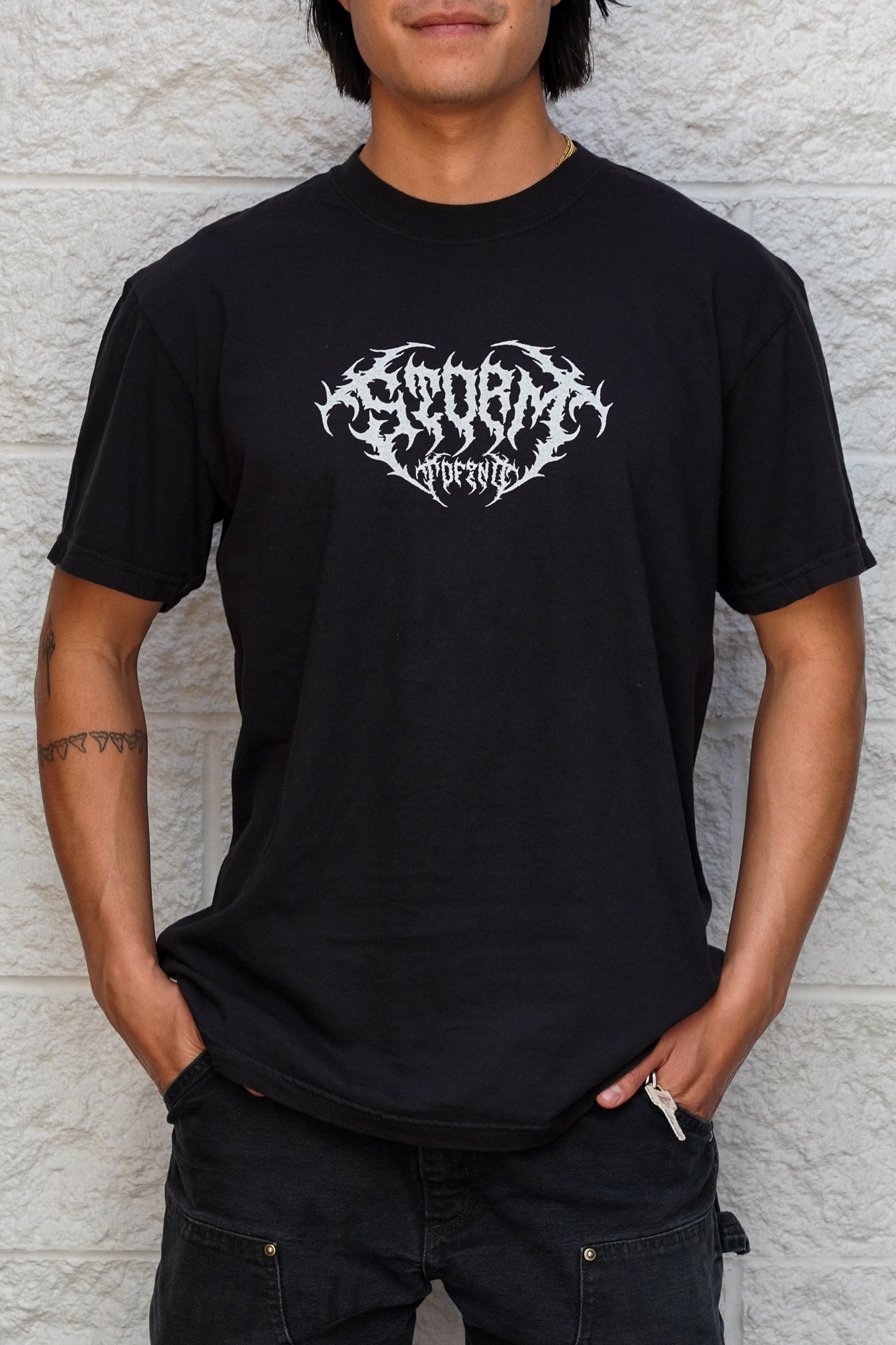 Person wearing a black t-shirt with a white logo against a textured wall.