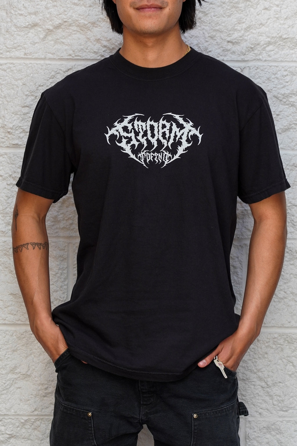 Person wearing a black t-shirt with a white logo against a textured wall.